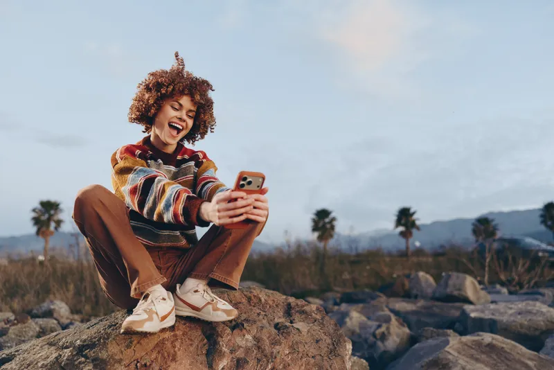 Woman outdoors lifestyle, laughing holding a smartphone, smile, sitting on rocky rocks, natural light, cloudy sky, palm trees, rocky coastline, breeze, fresh air, relaxed posture