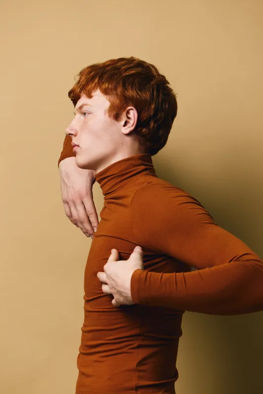 Young man with red hair in brown turtleneck posing sideways against beige background. Stylish fashion model showing profile with serious expression and hand gesture.
