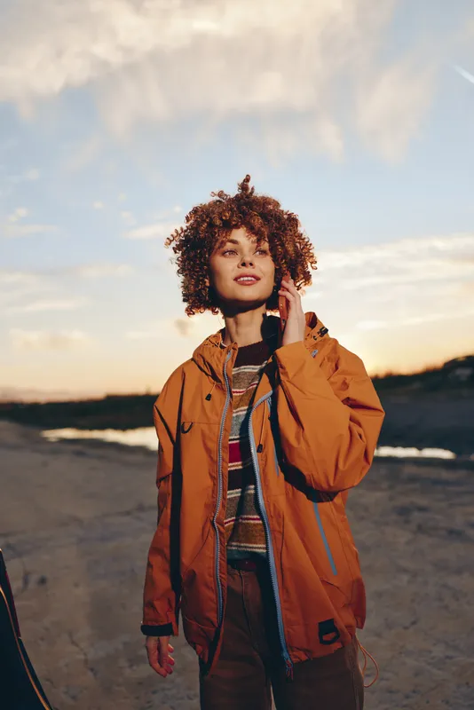 Woman outdoors at sunset on beach, curly hair blowing in light wind, wearing orange jacket and casual striped top, relaxed portrait pose, warm golden sky, soft light, lifestyle