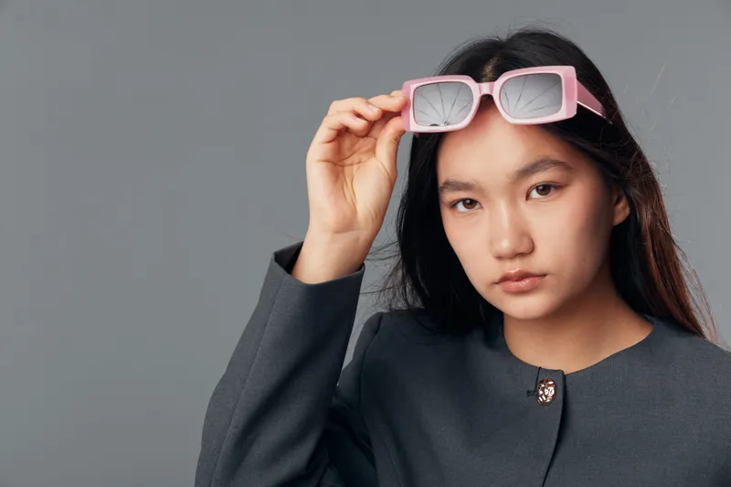 Young woman with long hair wearing dark blazer and holding pink sunglasses on gray background. Serious expression with natural makeup and casual modern style for portrait.