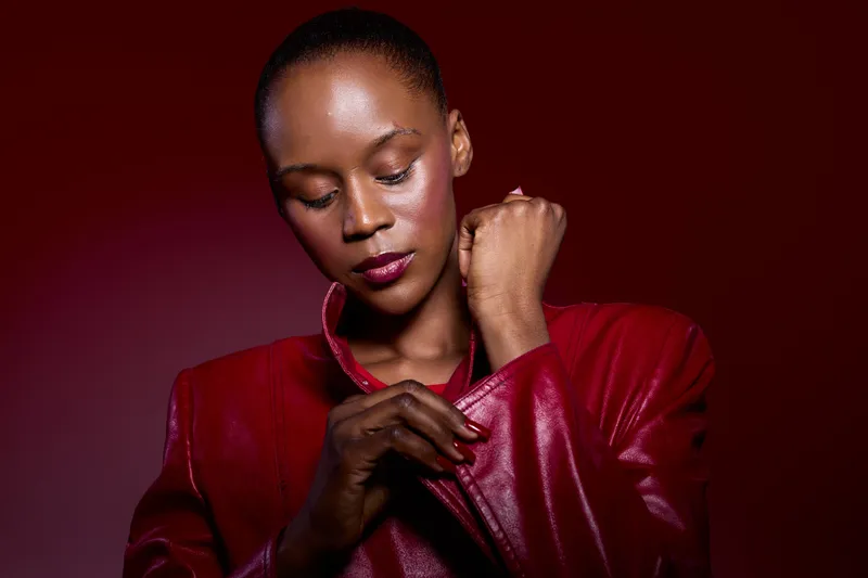Portrait of dark skinned African woman in red leather jacket adjusting sleeve. Studio shot with red background. Concept for fashion, beauty, confidence, and style in studio lighting.