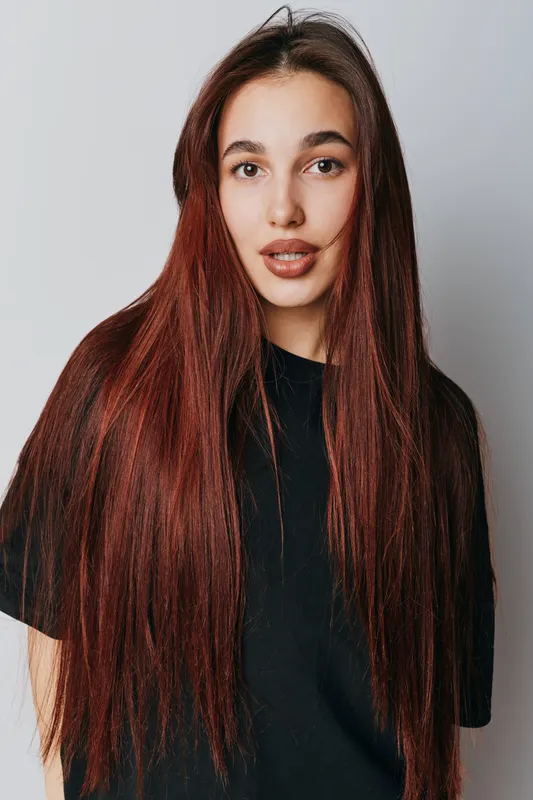 Young woman with long straight red hair wearing black t shirt posing with neutral expression on light background. Female model with natural makeup and casual style portrait.