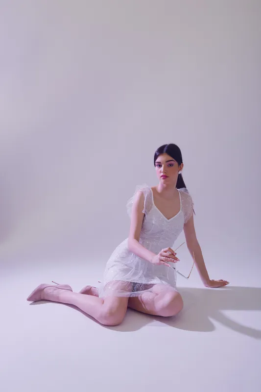 Young woman in a white dress kneels on the floor holding a pearl necklace. Studio shot with soft lighting. Concept of beauty, fashion, and elegance.