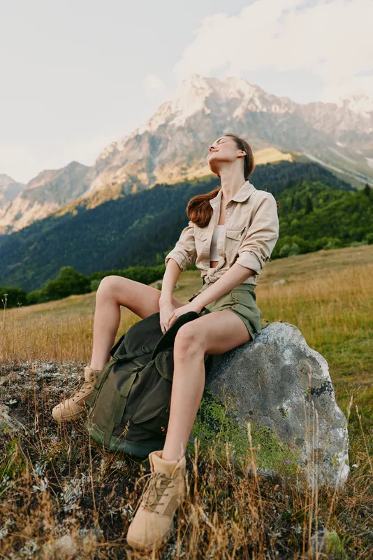A woman sits on a rock in a grassy meadow with mountains in the background, enjoying a peaceful outdoor moment. Concept for natural living, serenity, and outdoor lifestyle.