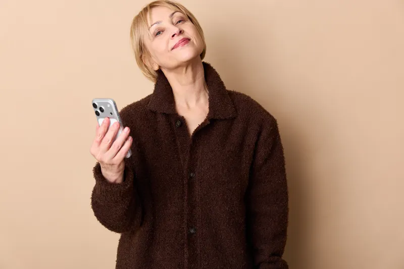 Woman with smartphone in hand, portrait on beige background. Communication and texting concept, modern lifestyle indoors, warm studio lighting, soft smile today