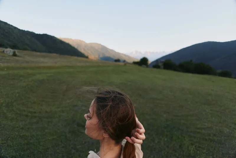 A young woman stands in a vast grassy field with distant mountains, her back to the camera as she gazes toward the horizon, bathed in soft evening light.