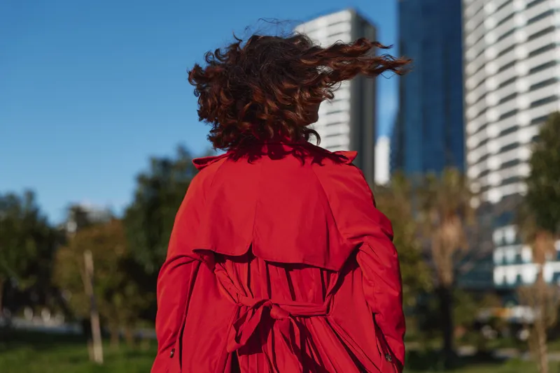 A young woman with curly hair wearing a bright red coat stands with her back to the camera in an urban park on a sunny day.