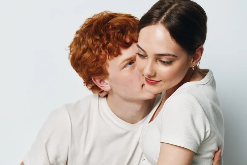 Couple hugging and intimate moment with young man kissing woman on cheek against white background. Love, affection and relationship with casual clothing and gentle expression.