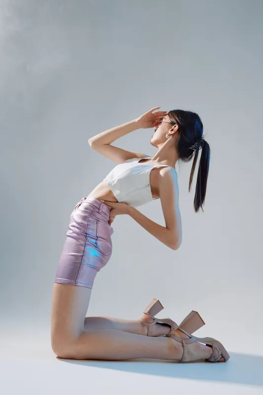 Slim woman in a white top and shiny skirt poses kneeling with hand on forehead. Studio shot on light background. Concept of beauty fashion and elegance.