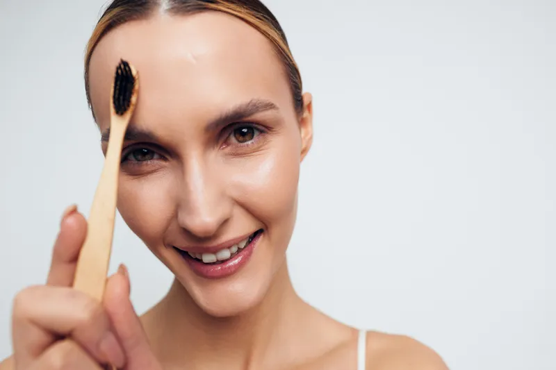 Young woman with blonde hair smiles gently while holding a bamboo toothbrush close to her face against a plain white background, promoting eco-friendly dental care.