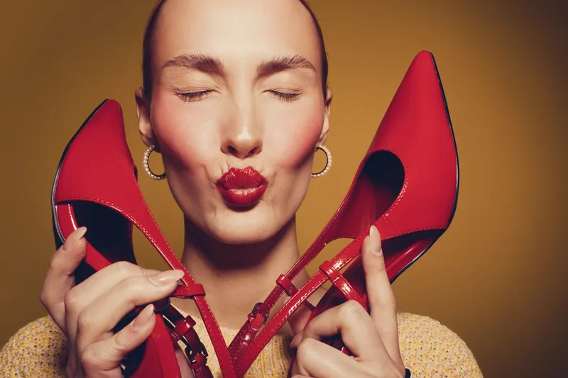 Woman with closed eyes holds red high heels near face, puckering lips. Studio portrait with warm lighting. Concept of fashion, style, beauty, and footwear accessories.