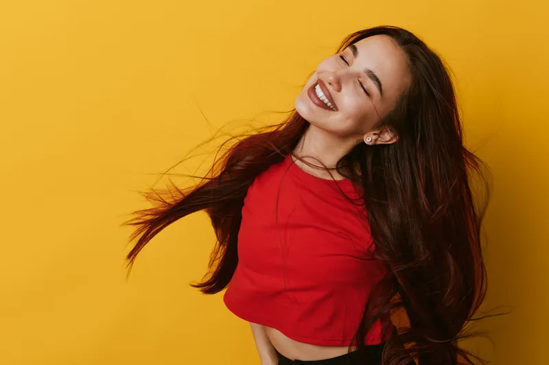 Young woman with long hair smiling and posing in red top against yellow background. Happy female model enjoying moment with closed eyes and joyful expression in studio.