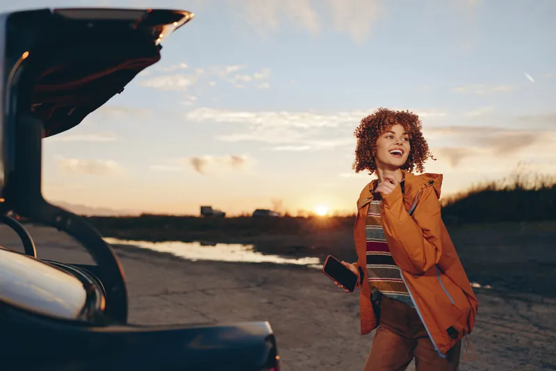 Woman outdoor lifestyle, walking on beach at sunset near a parked car, orange jacket and curly hair blowing, golden light and sea breeze, travel adventure mood, fresh air freedom