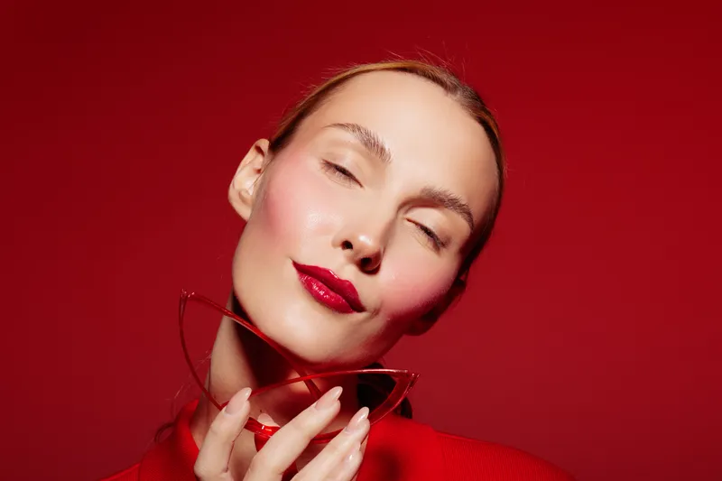 Close-up of a young woman with red lipstick and closed eyes holding red sunglasses on a red background. Concept of beauty, fashion, makeup, and elegance with vibrant colors.