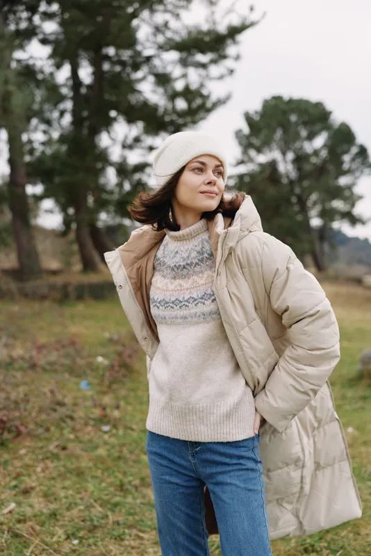 Woman in beige puffer coat standing in a park, wearing a knit sweater and beanie, enjoying a chilly day with trees and open field behind, calm and relaxed.