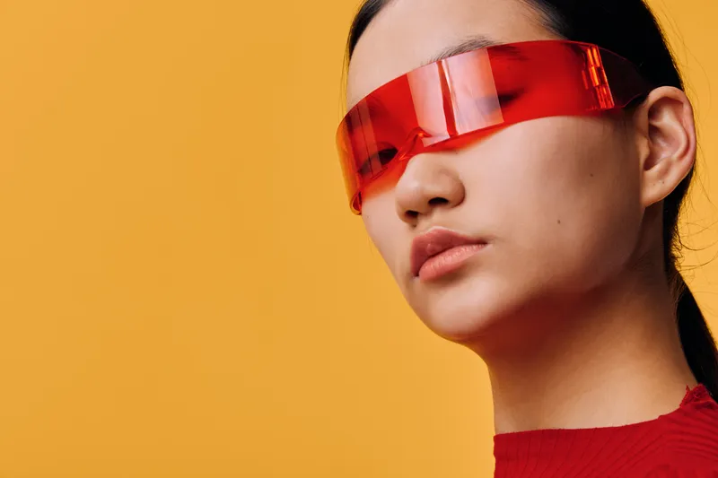Portrait of woman with long dark hair wearing futuristic red transparent glasses and red shirt against yellow background in studio. Fashion concept with minimalistic style and bright colors.