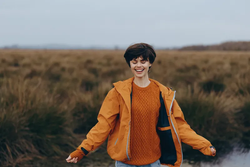 Woman smiling outdoors in a field, walking with orange jacket and sweater, countryside lifestyle in autumn, soft natural light, warm happy mood, nature landscape
