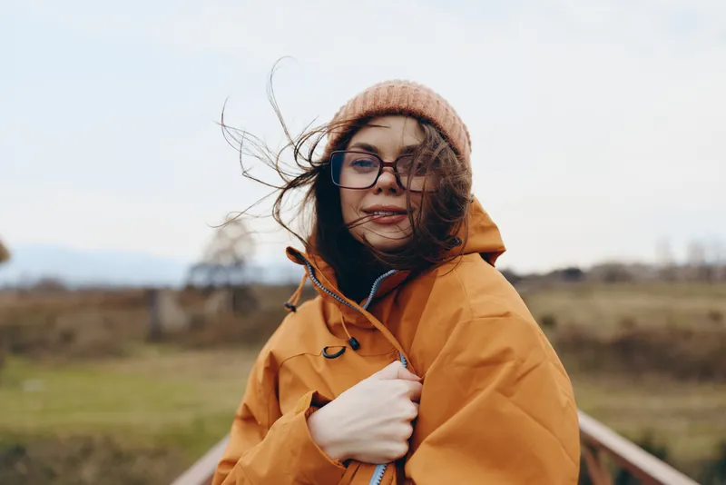 Woman lifestyle portrait outdoors in winter, wearing yellow coat, knitwear hat and glasses, wind moves hair as she adjusts jacket on rural road, natural light, calm countryside mood