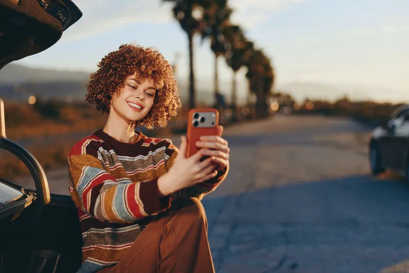 Woman lifestyle selfie with smartphone outdoors, smiling at sunset, curly hair and sweater, casual pose near road with bike, warm evening light, travel mood, golden hour portrait