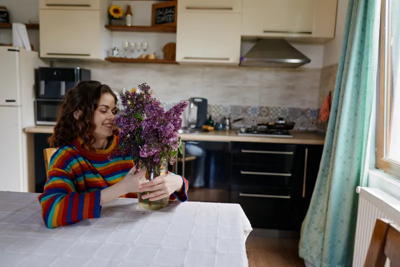 A young woman sits at a home kitchen table, admiring a bouquet of lilacs and smiling as sunlight fills the cozy room with a warm, inviting mood.