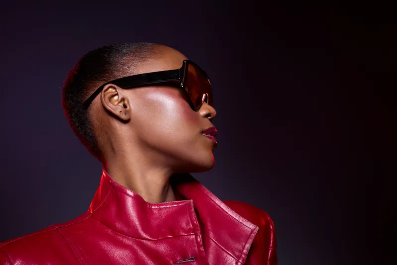 Profile of a confident black woman with dark skin wearing oversized sunglasses and a red leather jacket. Studio portrait with dramatic lighting on a dark purple background. Fashion concept.