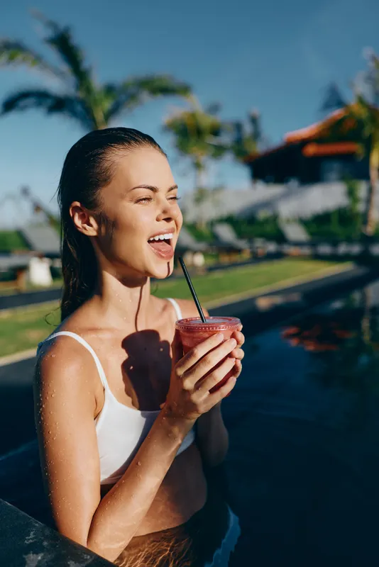 Young woman in white swimsuit enjoying a cold drink by the pool on a sunny day. Lifestyle concept with summer relaxation and tropical background. Happy mood with natural sunlight.