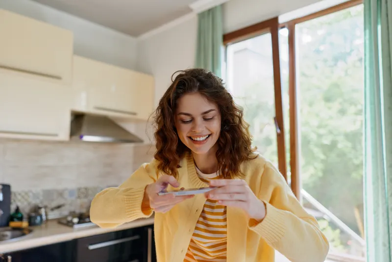 Smiling woman in a yellow cardigan using a smartphone in a bright kitchen, capturing home lifestyle with casual, cozy vibes and natural light