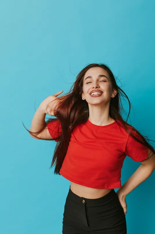 Young woman smiling with closed eyes and long hair wearing red shirt and black pants standing against blue background. Portrait of happy female enjoying moment with carefree expression.