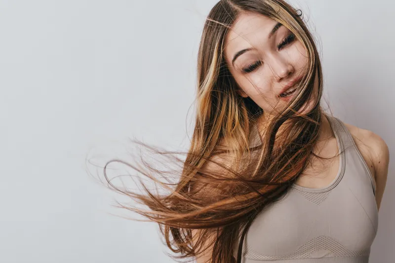 Young woman with long hair flowing, eyes closed wearing sleeveless top, posing naturally against plain background in casual style portrait with movement and serenity.