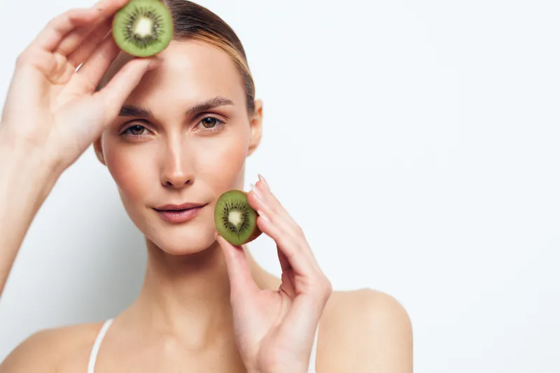 A focused woman holds fresh kiwis near her face in a bright minimalist studio, highlighting natural beauty, skincare confidence, healthy eating, and a lifestyle idea for wellness and daily self care.