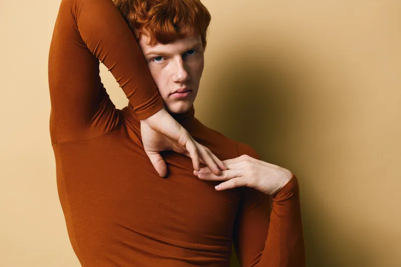 Young man with red hair and fair skin wearing fitted brown turtleneck sweater posing with arms raised against beige background in studio portrait.