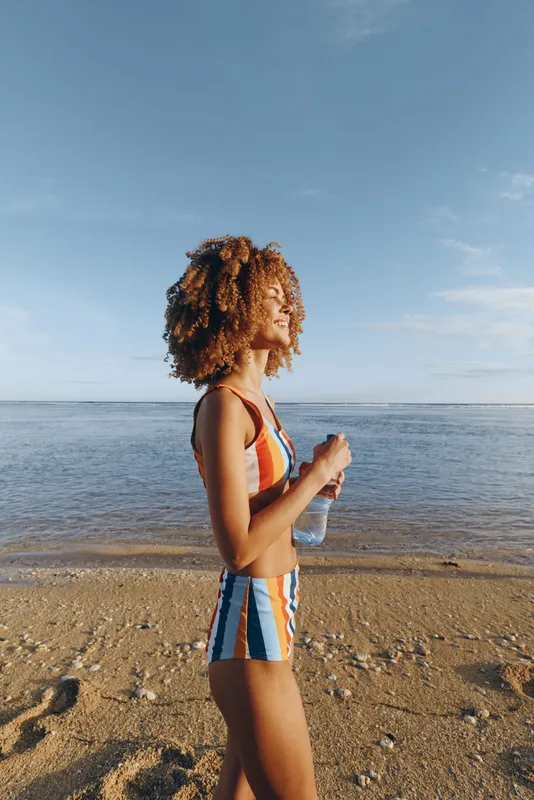 Young woman with curly hair walks along a sandy beach holding a water bottle while enjoying a sunny day by the calm sea, wearing a colorful striped swimsuit.