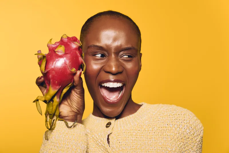 Smiling African woman with dark skin holds dragon fruit near her face, wearing beige sweater on yellow background. Concept of healthy lifestyle, exotic fruit, and natural beauty.