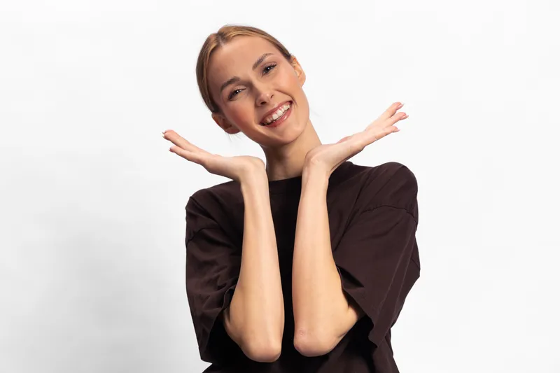 Smiling young woman in black shirt poses with hands near face, expressing happiness and joy. Studio portrait on white background with casual clothing and natural makeup.
