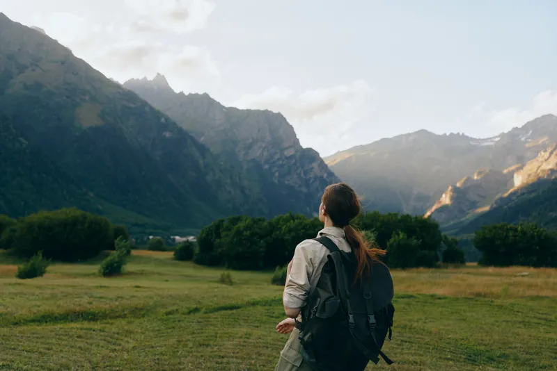 Young woman with backpack stands in a sunlit valley, gazing toward distant mountains, capturing a moment of exploration and calm amid nature.