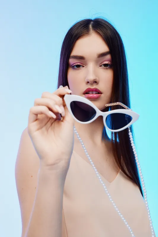 Young woman with long dark hair holds white sunglasses with pearl chain in front of blue background. Concept of beauty, fashion, and style accessory.