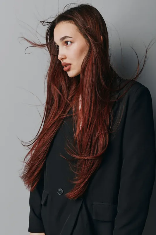 Young woman with long red hair wearing black blazer posing sideways against gray background. Natural look with loose hair strands and casual style in studio portrait photography.