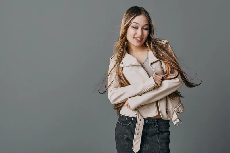 Young woman with long hair in casual jacket smiling and looking down on gray background. Beauty, fashion, lifestyle and relaxed pose with confidence and style.