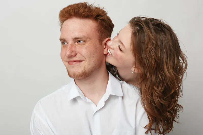 Young couple in white shirts share a tender moment, woman whispering in man’s ear. Studio portrait on light background. Concept of love, intimacy, and affection between partners.