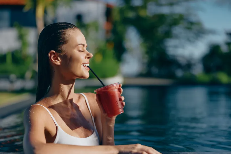 Smiling woman in a white swimsuit sips a red beverage beside a sunlit pool, enjoying a refreshing moment. Poolside lifestyle scene focused on relaxation, wellness, outdoor leisure by the water.