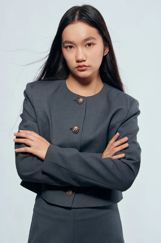 Woman with long black hair and serious expression in gray business suit standing with crossed arms against plain background. Confident female professional in formal outfit with button details.