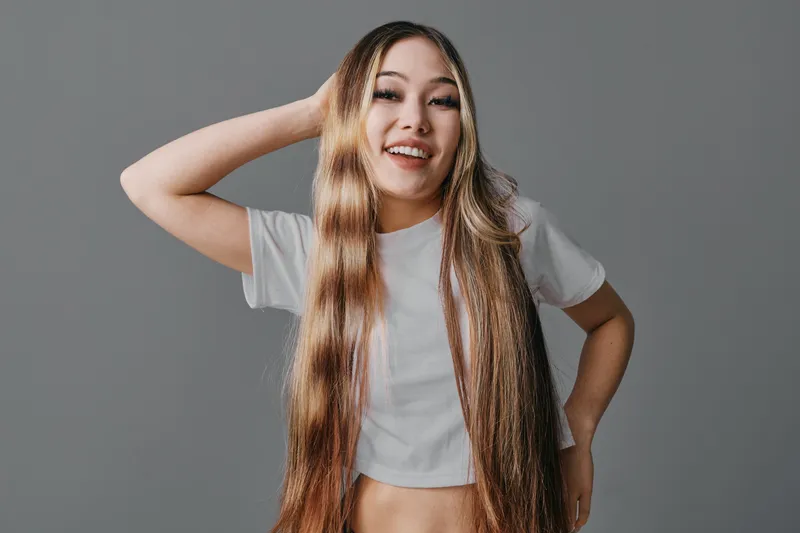 Young woman with long hair and white crop top smiling and posing with hand behind head on gray background. Happy model with casual style and confident attitude.
