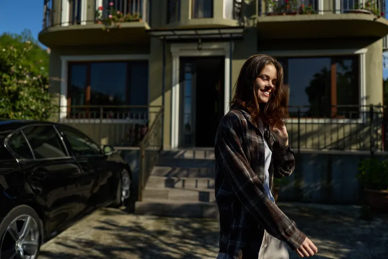 Young woman in a plaid shirt walks outdoors in front of a sunny building, turning toward the camera with a relaxed smile and casual confidence