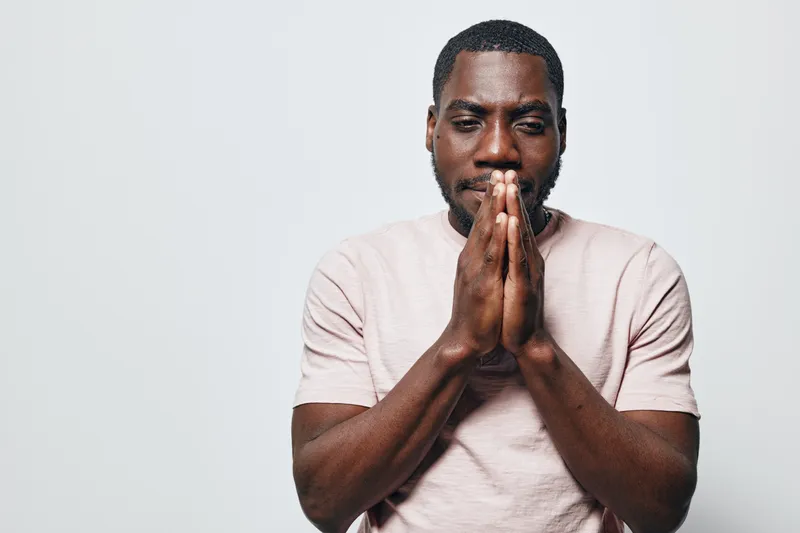 African American man with dark skin and beard standing with hands pressed together in prayer gesture, wearing light pink t shirt against white background. Thoughtful expression and casual style.