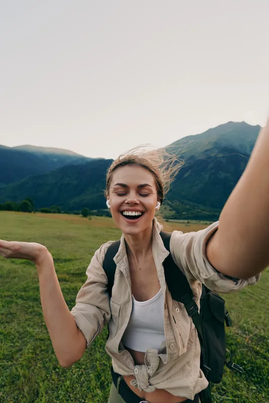 Smiling woman with backpack standing in a wide grassy field, mountains rising in the distance, sunlight warming her joyful expression, capturing a carefree outdoor lifestyle moment for travel,
