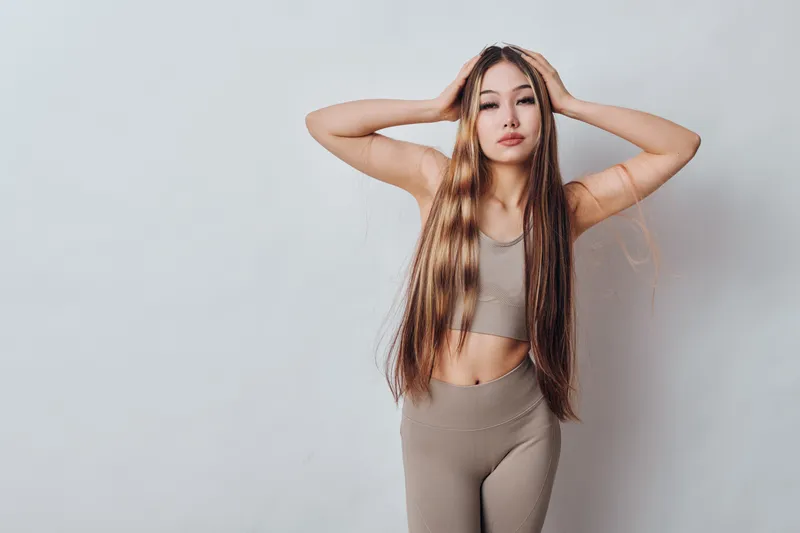 Young woman with long hair standing in casual sportswear with hands on head against grey background. Female model posing confidently in neutral toned outfit with fitness style.