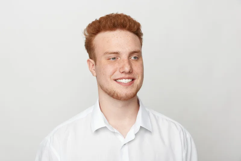 Smiling young man with red hair and beard wearing white shirt looks away on light background. Portrait of confident male model with casual style and positive expression in studio.