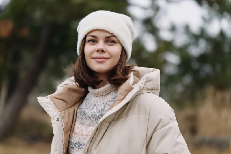 Outdoor portrait of a smiling woman in a beige jacket and knit sweater, wearing a white beanie, in a chilly park with soft natural light.