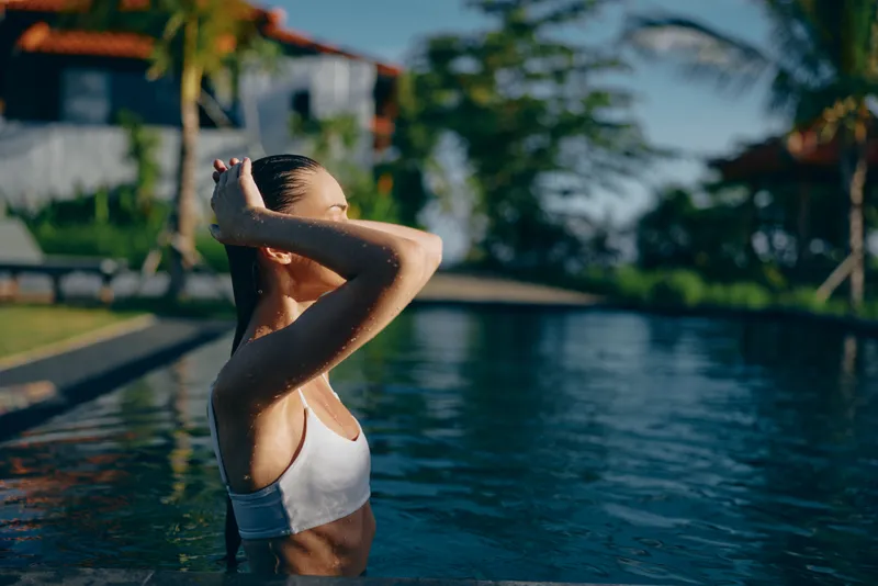 A woman in a white swim top stands in a sunlit pool, adjusting her hair. Tropical resort backdrop with palm trees and calm water. A lifestyle moment of leisure, wellness, and summer relaxation.