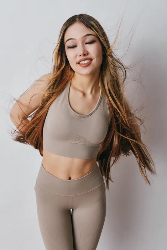 Young woman in beige fitness wear with long hair smiling and eyes closed standing on white background for wellness and healthy lifestyle concept.